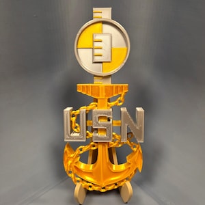 May include: A 3D-printed USN emblem in gold, silver, and white. The design includes an anchor, chain, and the letters "USN". The emblem is mounted on a small stand and features a circular element at the top.