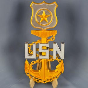 Puede incluir: Un emblema USN dorado y plateado, impreso en 3D, con un escudo con una estrella, las letras "USN" y un ancla. El emblema está sostenido por dos soportes de madera. El diseño incluye una cadena y se encuentra sobre un fondo gris.