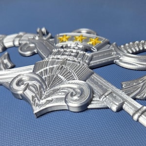 May include: Silver-colored military emblem featuring an anchor, sword, and decorative elements. The emblem has three gold stars at the top. The design is detailed and three-dimensional, set against a blue background.
