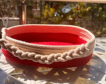 Puede incluir: Una cesta redonda, hecha a mano, con interior rojo y exterior beige y rojo. La cesta tiene un borde trenzado beige alrededor del borde superior. La cesta está sobre una mesa de mimbre blanca.
