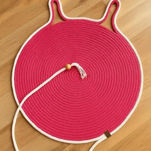 Op de afbeelding: Een ronde, felroze geweven mat met een witte touwrand en twee gebogen handvatten. Een wit touw loopt vanuit het midden en eindigt in een houten kraal en kwast. De mat ligt op een houten vloer.