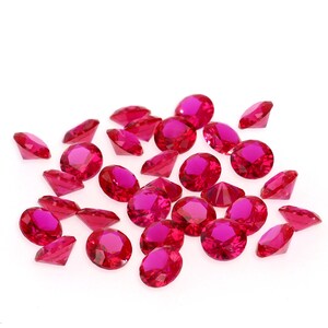 May include: A collection of vibrant, faceted, ruby-red gemstones scattered on a white surface. The gems vary in shape, with round and triangular cuts. The light catches the facets, creating a sparkling effect. Ideal for crafts or jewelry.