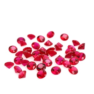 May include: A collection of small, faceted, red gemstones scattered on a white surface. The gems vary in shape, including round, oval, and triangular cuts. The gemstones are a vibrant shade of red, with a glossy finish.