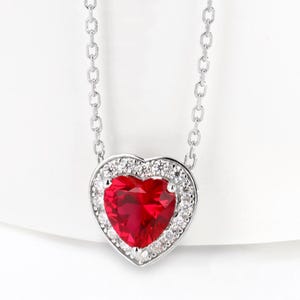 May include: A silver necklace featuring a heart-shaped, vibrant red gemstone pendant. The heart is surrounded by a halo of small, clear gemstones, and hangs from a delicate chain. The jewelry is set against a white background.