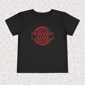 Peut inclure: T-shirt noir pour enfant avec un motif rouge. Le motif est une estampille circulaire avec le texte "CERTIFIED MISCHIEF MAKER CERTIFIED". Le t-shirt est en matière douce.