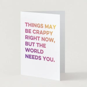 May include: A white greeting card with the text "THINGS MAY BE CRAPPY RIGHT NOW, BUT THE WORLD NEEDS YOU." The text is in a gradient of colors, from yellow to purple. The card is folded and stands upright.