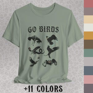 May include: Sage green t-shirt with the text "GO BIRDS" in a gothic font above a graphic of various black birds. The phrase "+11 COLORS" is printed at the bottom of the shirt.