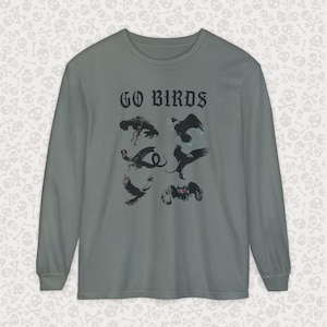 May include: A long-sleeve shirt in a muted green color. The shirt features the text "GO BIRDS" in a gothic font above a graphic of several stylized birds in black. The birds are in various poses, some with outstretched wings.