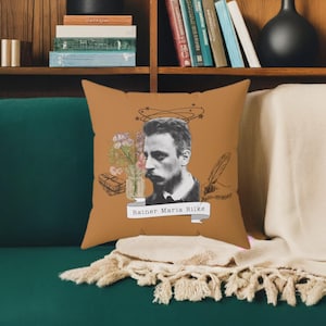 Può includere: Un cuscino marrone per divano con un ritratto di Rainer Maria Rilke, un mazzo di fiori e una penna. Il cuscino ha il testo "Rainer Maria Rilke".