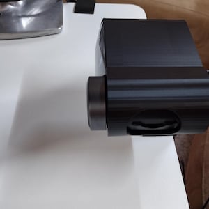 May include: A black electronic device with a cylindrical lens and a rounded body. The device is connected to a black cable. It is placed on a white surface, with a silver object visible in the background.