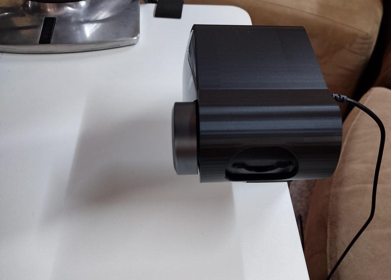 May include: A black electronic device with a cylindrical lens and a rounded body. The device is connected to a black cable. It is placed on a white surface, with a silver object visible in the background.