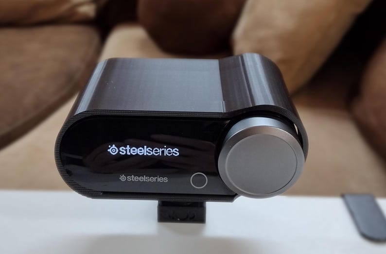 May include: A black and silver SteelSeries audio device with a large control knob. The device has the SteelSeries logo and text on the front. The device is sitting on a white surface.