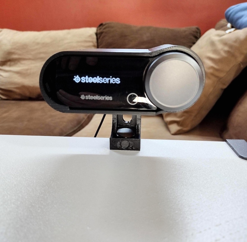 May include: A black SteelSeries audio device with a silver dial and a black stand. The device has the SteelSeries logo printed on the front. The device is mounted on a white surface.