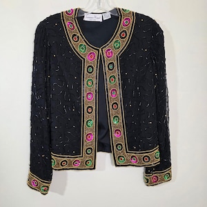 May include: Black beaded jacket with colorful circular accents. The jacket features a V-neck design and is adorned with gold, green, pink, and red embellishments. The sleeves and body are covered in small beads. The jacket is hanging on a white hanger.