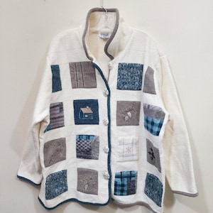 May include: Cream-colored quilted jacket with square patchwork design. The jacket features embroidered squares with house, pinecone, and snowflake motifs. It has a button-up front and a gray trim.