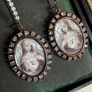 May include: A pair of vintage-style earrings. Each earring features an oval pendant with a sepia-toned image of a religious figure, surrounded by a halo of clear crystals. The earrings have a bronze-colored metal frame and a decorative hook.