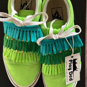 May include: Green canvas Vans sneakers with multi-colored fringe and gold accents. The shoes have white laces and a white tag that says "Play Ded one of a kind hand crafted - made in the USA".
