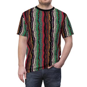 Op de afbeelding: Een t-shirt met korte mouwen met een zwarte kraag en een kleurrijk, verticaal gestreept patroon. De strepen omvatten rood, groen, zwart, wit en paars, waardoor een gestructureerd, golvend ontwerp ontstaat. Het shirt wordt gedragen met een blauwe spijkerbroek.