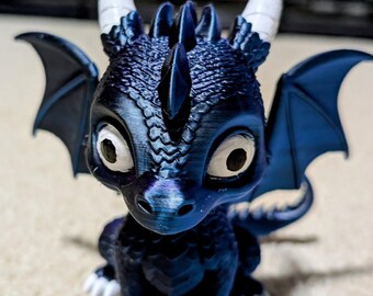 May include: A blue dragon figurine with white horns and claws. The dragon has large, expressive eyes and small, dark blue wings. The figurine is detailed with textured scales and a spiky spine, giving it a whimsical, fantasy aesthetic.