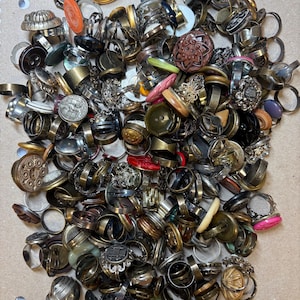 Puede incluir: Una colección de anillos variados, con diversos diseños y materiales. Los anillos incluyen tonos plateados, dorados y mixtos, con adornos como botones, piedras y elementos decorativos. Los anillos son de diferentes tamaños y estilos.