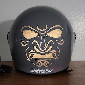 May include: A dark gray motorcycle helmet with a gold-colored samurai mask design on the front. The text "Shift to Six" is printed at the bottom. The helmet is sitting on a wooden surface.