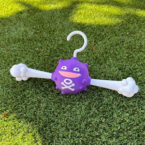 Puede incluir: Una percha morada novedosa con una cara de dibujos animados y un diseño de calavera y huesos cruzados. La percha tiene brazos blancos con extremos en forma de hueso. El gancho es blanco y la percha descansa sobre hierba verde.