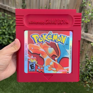 May include: A red Game Boy cartridge featuring the Pokémon logo and a Charizard illustration. The cartridge has the text "Nintendo GAME BOY" and "THIS SIDE OUT". The game is rated "Everyone" by the ESRB.