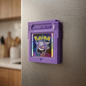 May include: A purple Game Boy cartridge magnet featuring the Pok&eacute;mon Gengar. The magnet has the Nintendo Game Boy logo and the Pok&eacute;mon logo. The cartridge is attached to a stainless steel surface.