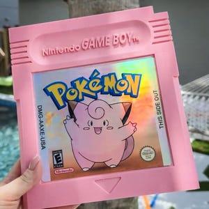 Puede incluir: Un cartucho rosa de Nintendo Game Boy con el juego Pokémon. El cartucho tiene el texto "Nintendo GAME BOY" en la parte superior y el logotipo de Pokémon con un personaje Clefairy rosa. También es visible el texto "THIS SIDE OUT".