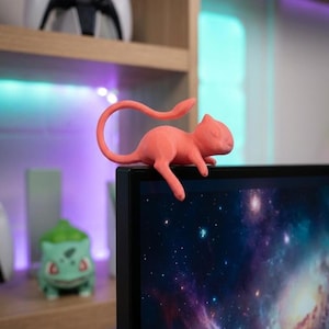 Puede incluir: Figura de color coral de una criatura felina, apoyada en el borde de un monitor de ordenador. La figura tiene una larga cola y está colocada sobre un fondo de imagen de galaxia en la pantalla. Un mando de videojuegos es visible en una estantería al fondo.