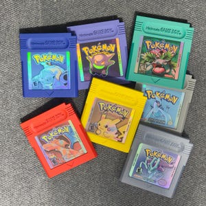 Puede incluir: Una colección de cartuchos de Pokemon Game Boy de colores. Los cartuchos son de varios colores, incluyendo rojo, amarillo, azul, morado y verde. Cada cartucho presenta una etiqueta holográfica con personajes de Pokemon y el logotipo "Pokemon".