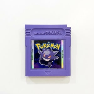 May include: A purple Nintendo Game Boy cartridge featuring the Pok&eacute;mon game. The label displays the Pok&eacute;mon logo and a grinning Gengar character. The cartridge has the text "Nintendo GAME BOY" at the top.