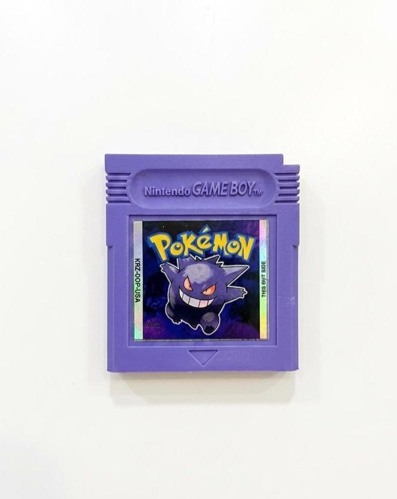 May include: A purple Nintendo Game Boy cartridge featuring the Pok&eacute;mon game. The label displays the Pok&eacute;mon logo and a grinning Gengar character. The cartridge has the text "Nintendo GAME BOY" at the top.