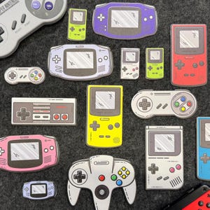 Puede incluir: Una colección de pegatinas coloridas troqueladas de consolas y mandos de videojuegos. Las pegatinas presentan diseños de consolas portátiles clásicas, mandos y un mando de Super Nintendo. Los colores incluyen morado, rosa, amarillo, verde, rojo y gris.