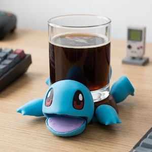 May include: A light blue turtle-shaped coaster with a smooth finish, large dark eyes, and an open mouth showing a purple interior. The coaster supports a clear glass filled with a dark brown liquid, featuring four flippers extending outward.