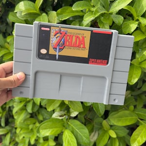 Puede incluir: Un cartucho de juego gris de Super Nintendo para "The Legend of Zelda: A Link to the Past". La etiqueta presenta el título y el logotipo del juego, con un diseño dorado, rojo y azul. El cartucho se sostiene contra un fondo de follaje verde.