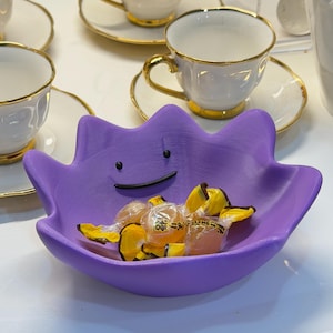 May include: A purple bowl shaped like the character Ditto, featuring a simple black line for a mouth and two black dots for eyes. The bowl is filled with several small, individually wrapped yellow candies with clear plastic packaging.