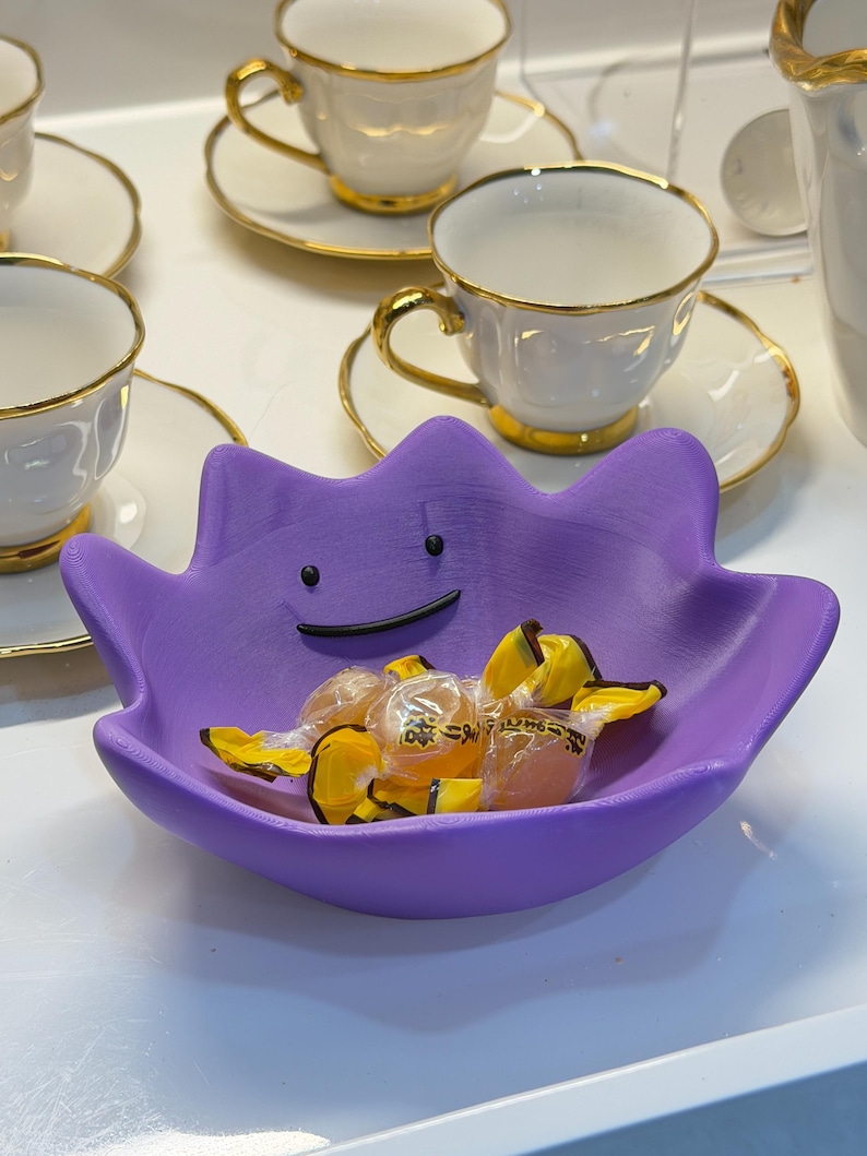 May include: A purple bowl shaped like the character Ditto, featuring a simple black line for a mouth and two black dots for eyes. The bowl is filled with several small, individually wrapped yellow candies with clear plastic packaging.