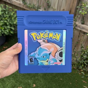 May include: A blue Nintendo Game Boy cartridge featuring the Pokémon logo and a Blastoise illustration. The cartridge has the text "Nintendo GAME BOY" and "THIS SIDE OUT". The game is rated "Everyone" by the ESRB.