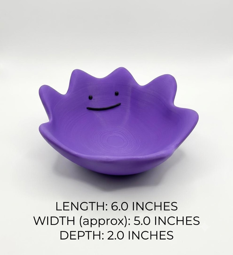 May include: A purple bowl shaped like a character with a wavy, irregular rim and a simple black line for a mouth and two black dots for eyes. The bowl has a length of 6.0 inches, a width of approximately 5.0 inches, and a depth of 2.0 inches.