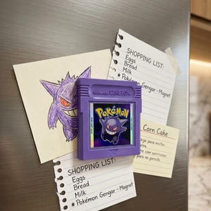 May include: A purple Nintendo Game Boy cartridge magnet featuring the Pok&eacute;mon Gengar, alongside handwritten shopping lists and a drawing of Gengar. The lists include items like eggs, bread, and milk.