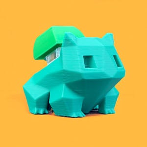 May include: A teal 3D-printed figure of a Bulbasaur, a popular character from the Pokémon franchise, with a green bulb on its back. The figure has a geometric, polygonal design and is set against a bright orange background.