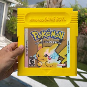 Pode incluir: Um cartucho amarelo de Game Boy com o jogo Pokémon Special Pikachu Edition. O cartucho tem o logotipo da Nintendo e o título do jogo em destaque. A arte da capa do jogo mostra Pikachu. O cartucho é segurado em uma mão.