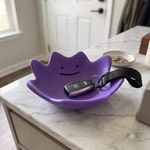 Può includere: Un vassoio viola a forma di ciotola con il volto di un personaggio caratterizzato da due piccoli punti neri per gli occhi e una sottile linea nera curva per la bocca. Una chiave dell'auto nera con un cinturino in tessuto nero poggia nel vassoio.