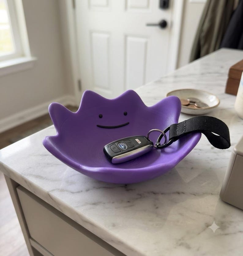 May include: A purple, bowl-shaped tray with a character face featuring two small black dots for eyes and a thin, curved black line for a mouth. A black car key fob with a black fabric strap is resting inside the bowl-shaped tray.