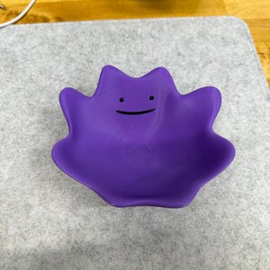 May include: A purple bowl shaped like the character Ditto, featuring a smooth, rounded form with irregular, wavy edges. The center of the bowl displays a simple, minimalist face consisting of two small black dots for eyes and a thin, curved black line for a mouth.