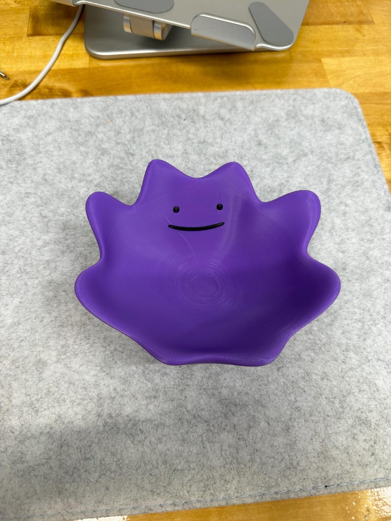 May include: A purple bowl shaped like the character Ditto, featuring a smooth, rounded form with irregular, wavy edges. The center of the bowl displays a simple, minimalist face consisting of two small black dots for eyes and a thin, curved black line for a mouth.