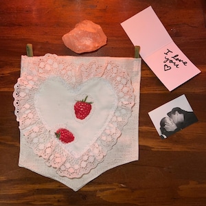 Könnte beinhalten: Ein weißes herzförmiges Kissen mit rosa Spitzenbesatz und gestickten roten Himbeeren. Das Kissen hängt an einer Wand mit einem handgeschriebenen Zettel, auf dem "Ich liebe dich" steht, und einem kleinen rosa Stein.