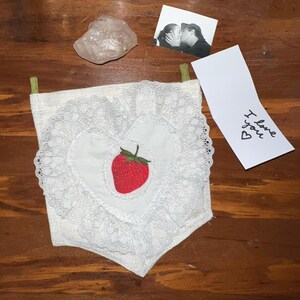 Peut inclure: Une pochette en tissu blanc en forme de cœur avec une fraise brodée rouge et une bordure en dentelle. La pochette est suspendue à un ruban vert. Une carte blanche avec le texte "Je t'aime" est en arrière-plan.