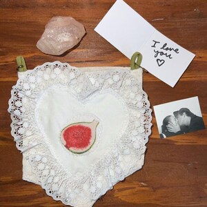 Puede incluir: Una almohada blanca en forma de corazón con un borde de encaje y un higo rojo bordado en el centro. La almohada está colgada en una pared de madera con un cristal rosa y una tarjeta que dice "Te amo" en ella.